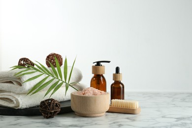 Photo of Spa treatment. Towels, sea salt, brush, bottles of cosmetic products and palm leaf on white marble table, space for text