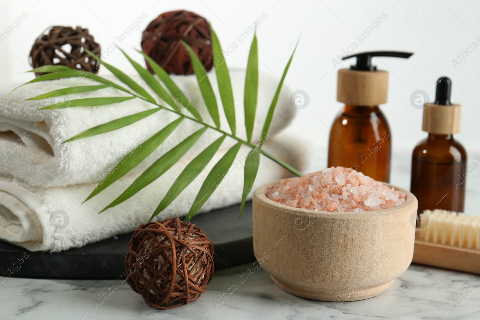 Photo of Spa treatment. Towels, sea salt, brush, bottles of cosmetic products and palm leaf on white marble table, closeup