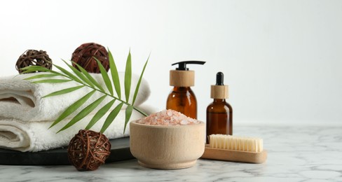 Photo of Spa treatment. Towels, sea salt, brush, bottles of cosmetic products and palm leaf on white marble table, space for text