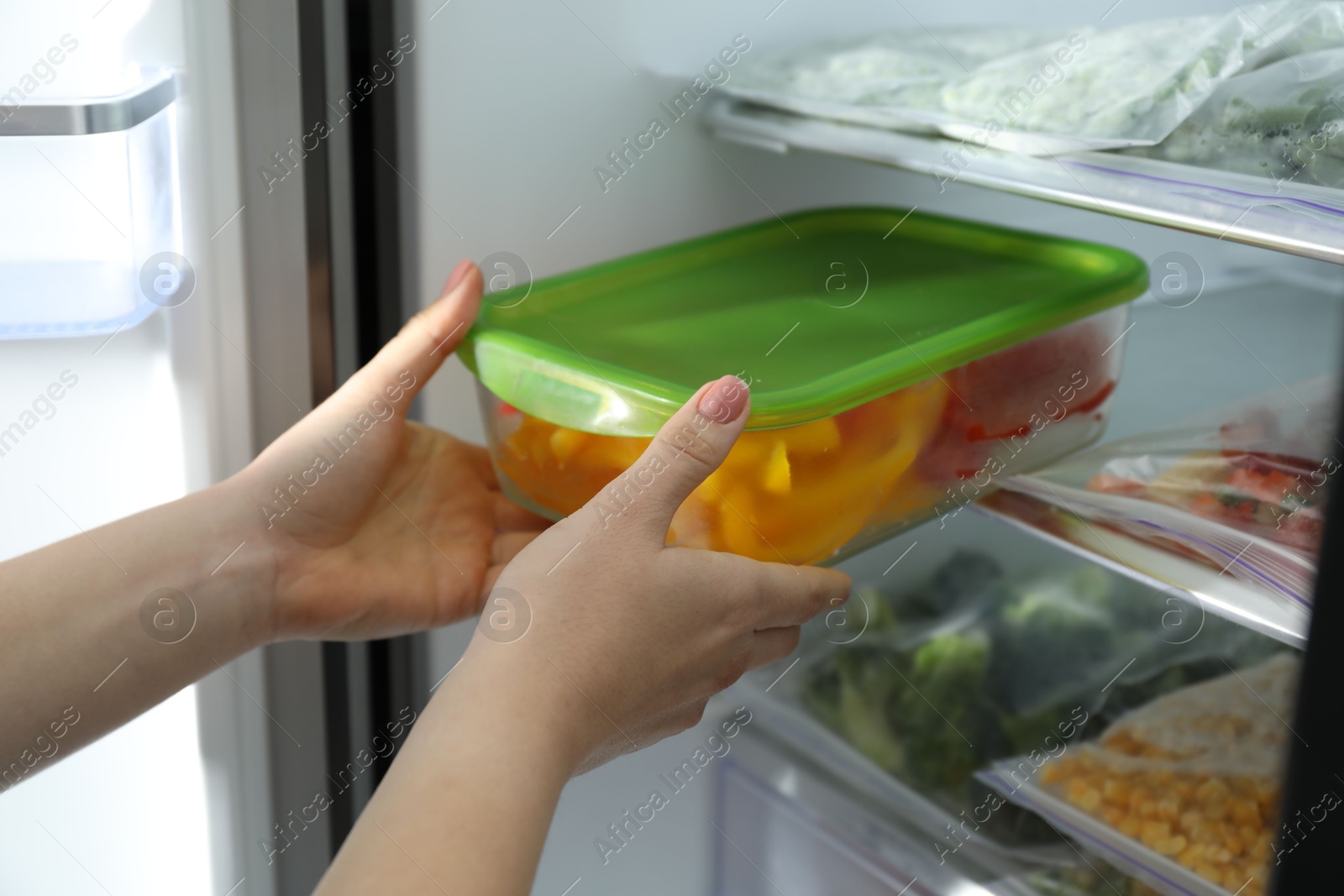 Photo of Woman taking glass container with frozen cut bell pepper from refrigerator, closeup