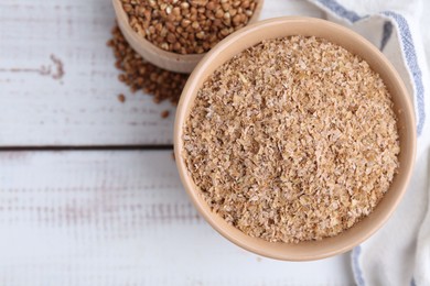 Photo of Buckwheat bran in bowl and grains on white wooden table, top view. Space for text