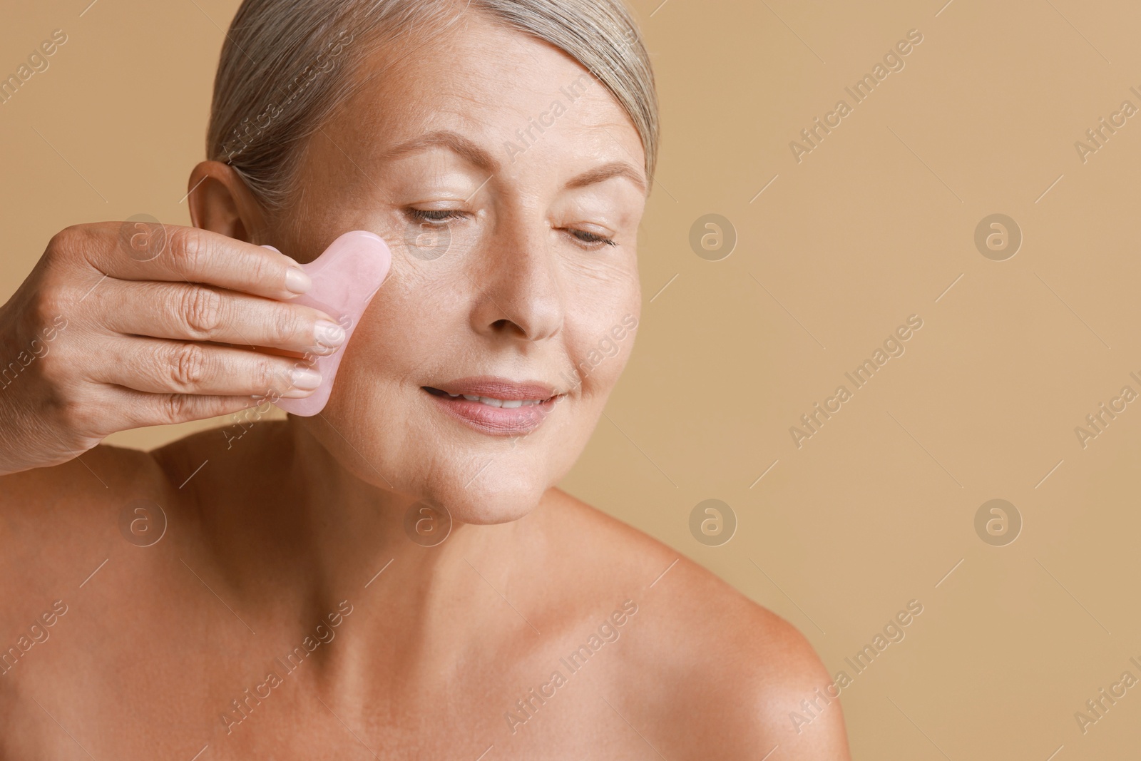 Photo of Beautiful woman doing facial massage with gua sha tool on beige background, space for text