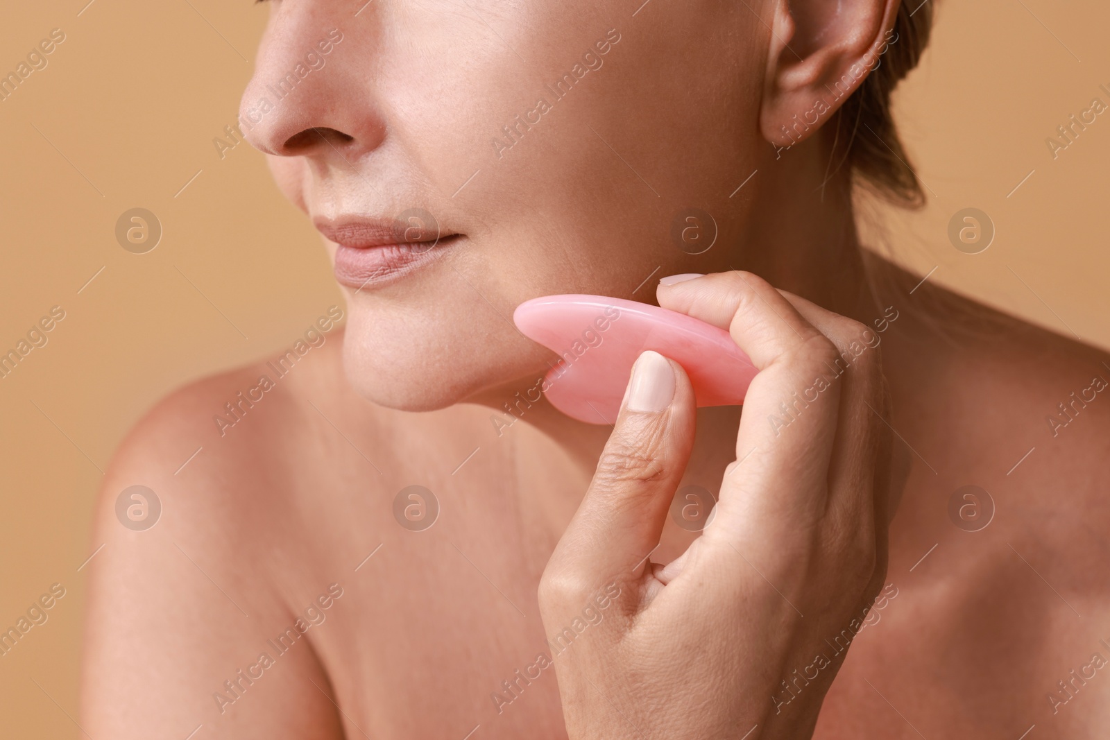 Photo of Woman doing facial massage with gua sha tool on beige background, closeup