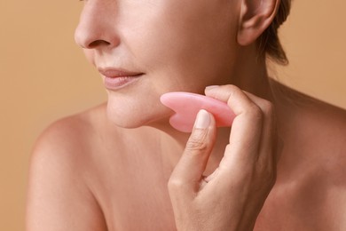 Woman doing facial massage with gua sha tool on beige background, closeup