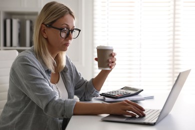 Photo of Budget planning. Woman with paper cup of coffee using laptop at table indoors