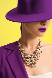 Fashionable woman with purple lipstick, hat and jacket on yellow background