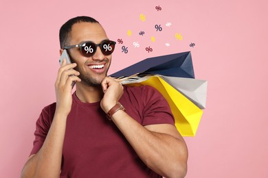 Man with sunglasses and shopping bags talking on phone against pink background. Percent signs on lenses. Discount, hot offer