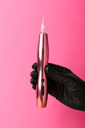 Photo of Professional cosmetologist holding permanent makeup machine on pink background, closeup