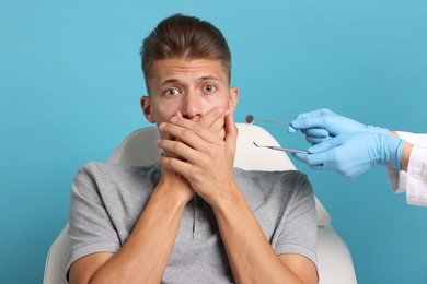 Dental phobia. Dentist working with scared man on light blue background, closeup