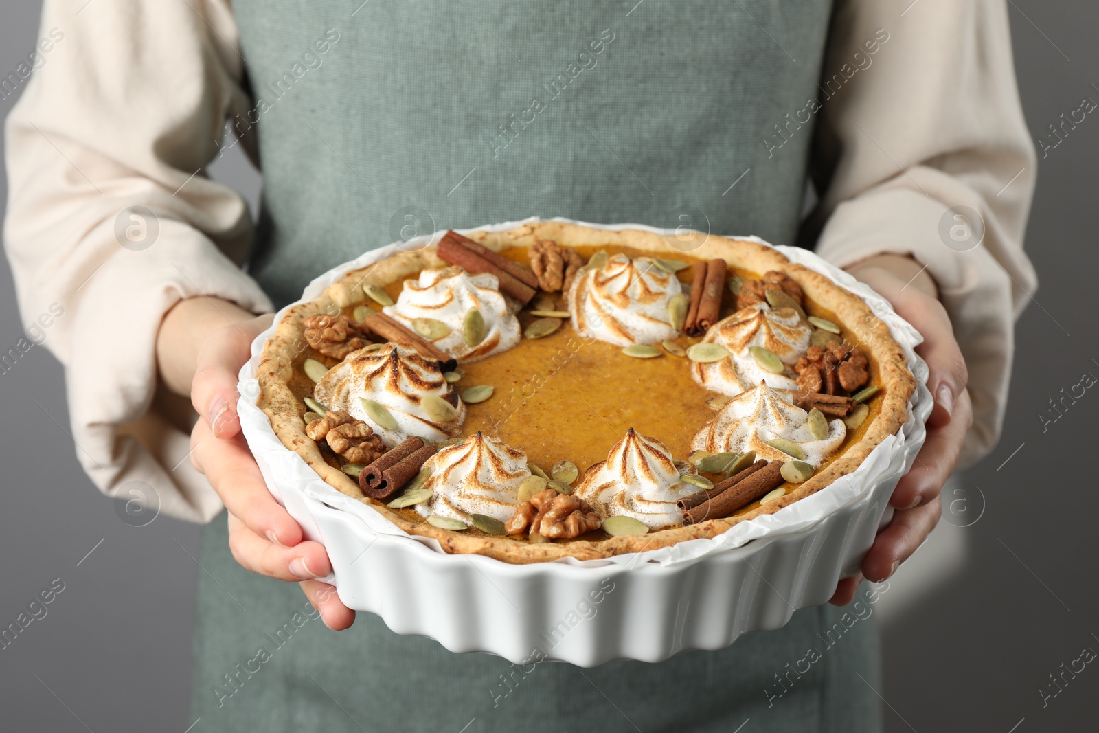 Photo of Woman holding homemade pumpkin pie with whipped cream, seeds and cinnamon on grey background, closeup