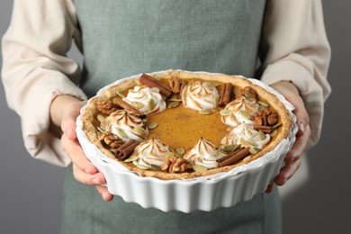 Woman holding homemade pumpkin pie with whipped cream, seeds and cinnamon on grey background, closeup