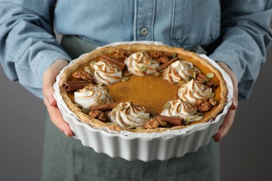 Woman holding homemade pumpkin pie with whipped cream, seeds and cinnamon on grey background, closeup