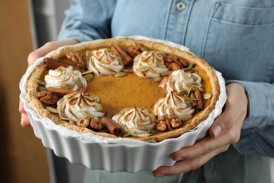 Photo of Woman holding homemade pumpkin pie with whipped cream, seeds and cinnamon on blurred background, closeup