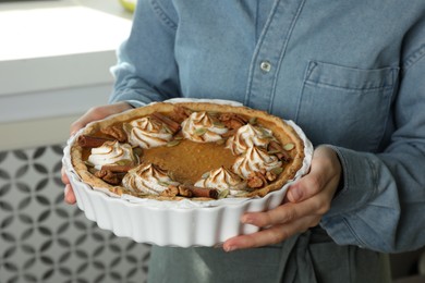 Woman holding homemade pumpkin pie with whipped cream, seeds and cinnamon on blurred background, closeup