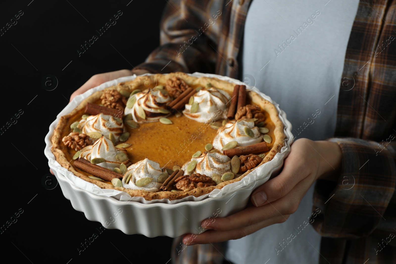 Photo of Woman holding homemade pumpkin pie with whipped cream, seeds and cinnamon on black background, closeup