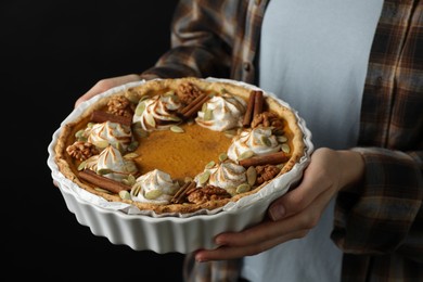 Woman holding homemade pumpkin pie with whipped cream, seeds and cinnamon on black background, closeup