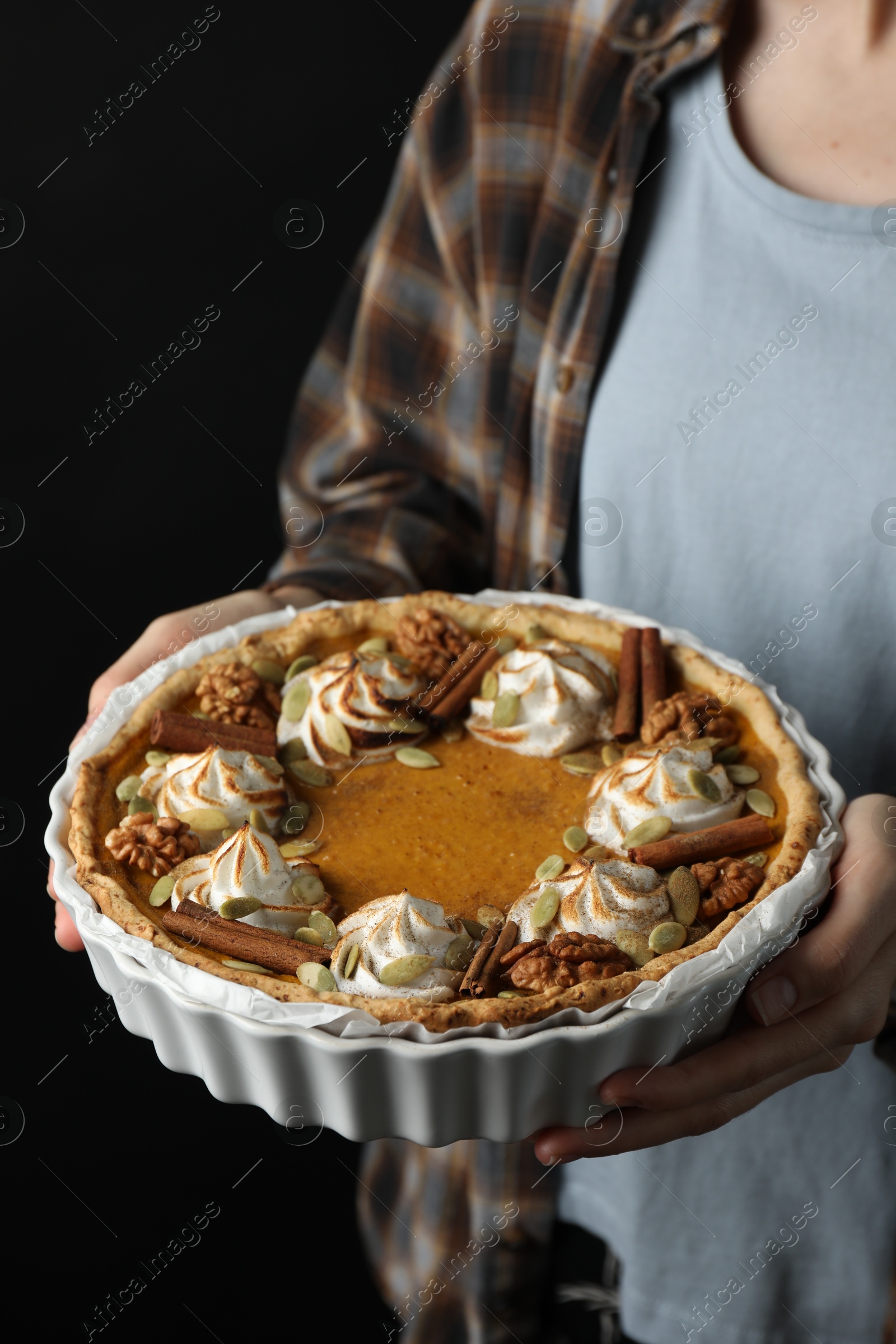 Photo of Woman holding homemade pumpkin pie with whipped cream, seeds and cinnamon on black background, closeup