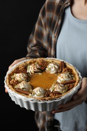 Woman holding homemade pumpkin pie with whipped cream, seeds and cinnamon on black background, closeup