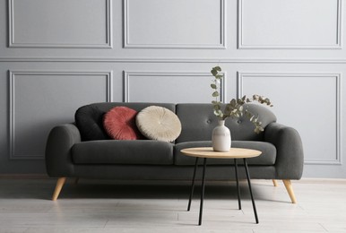 Photo of Stylish sofa with cushions and eucalyptus branches in vase on coffee table in room