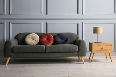 Stylish sofa with cushions and lamp on side table near grey wall