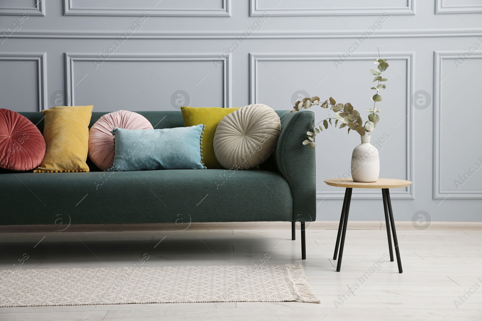 Photo of Stylish sofa with cushions and eucalyptus branches in vase on side table near grey wall