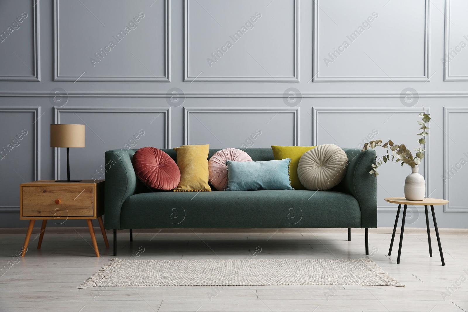 Photo of Stylish sofa with cushions, lamp and eucalyptus branches on side tables near grey wall