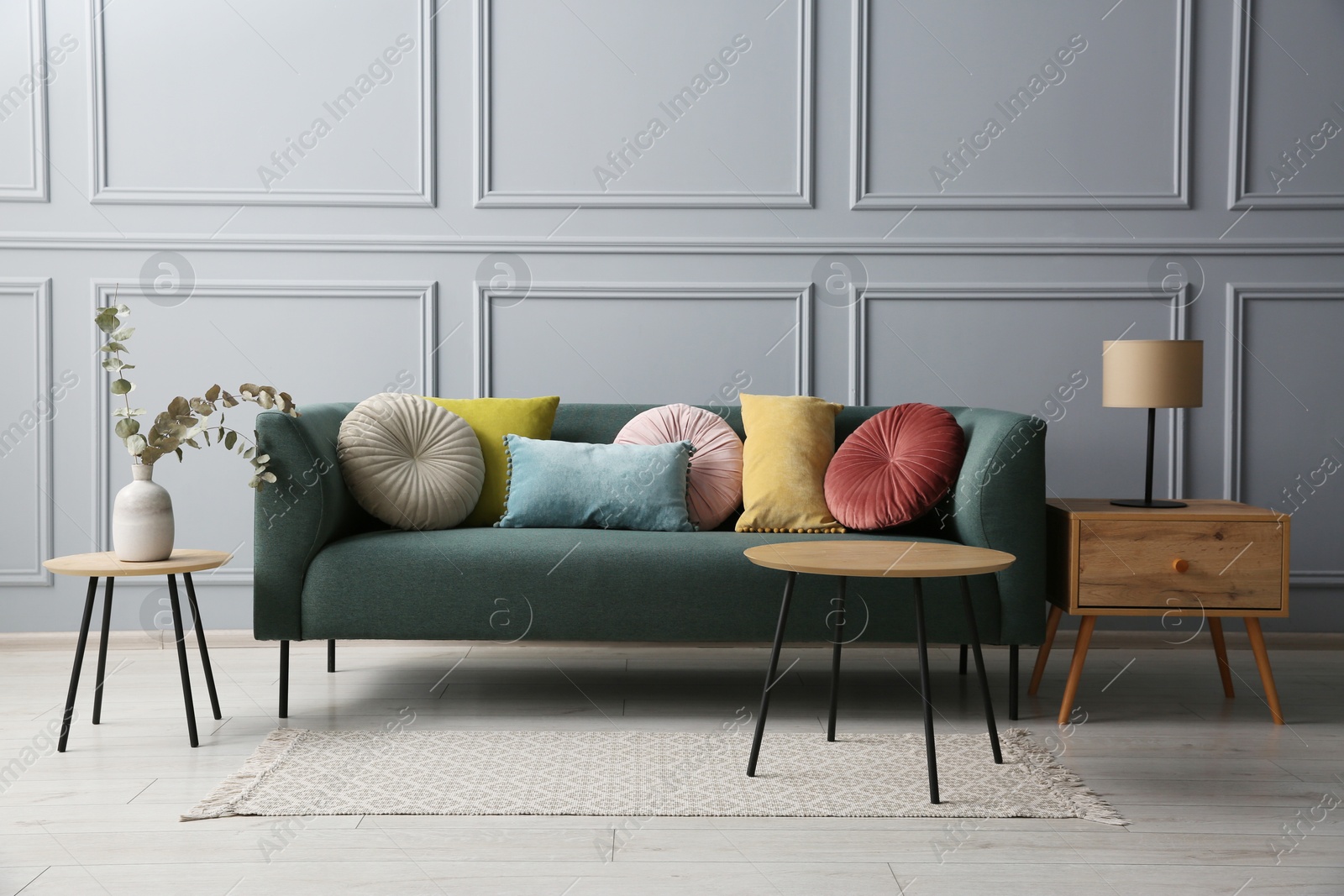 Photo of Stylish sofa with cushions, lamp and eucalyptus branches on side tables in room