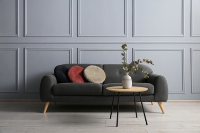 Photo of Stylish sofa with cushions and eucalyptus branches in vase on coffee table in room