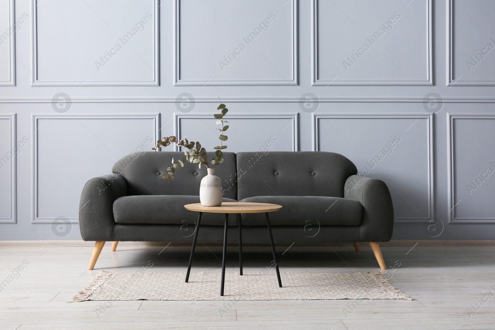 Photo of Stylish sofa and eucalyptus branches in vase on coffee table near grey wall