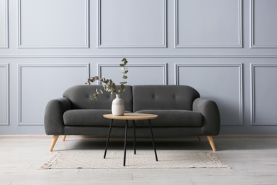 Photo of Stylish sofa and eucalyptus branches in vase on coffee table near grey wall