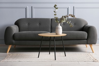 Photo of Stylish sofa and eucalyptus branches in vase on coffee table near grey wall