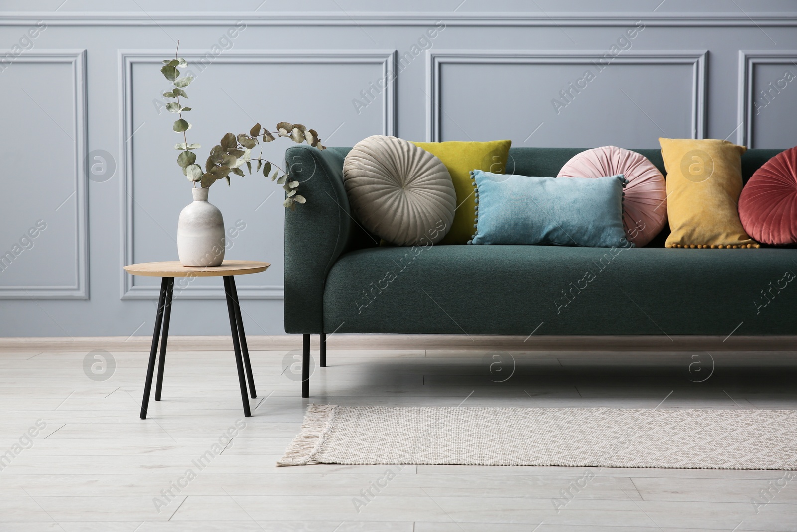 Photo of Stylish sofa with cushions and eucalyptus branches in vase on side table near grey wall