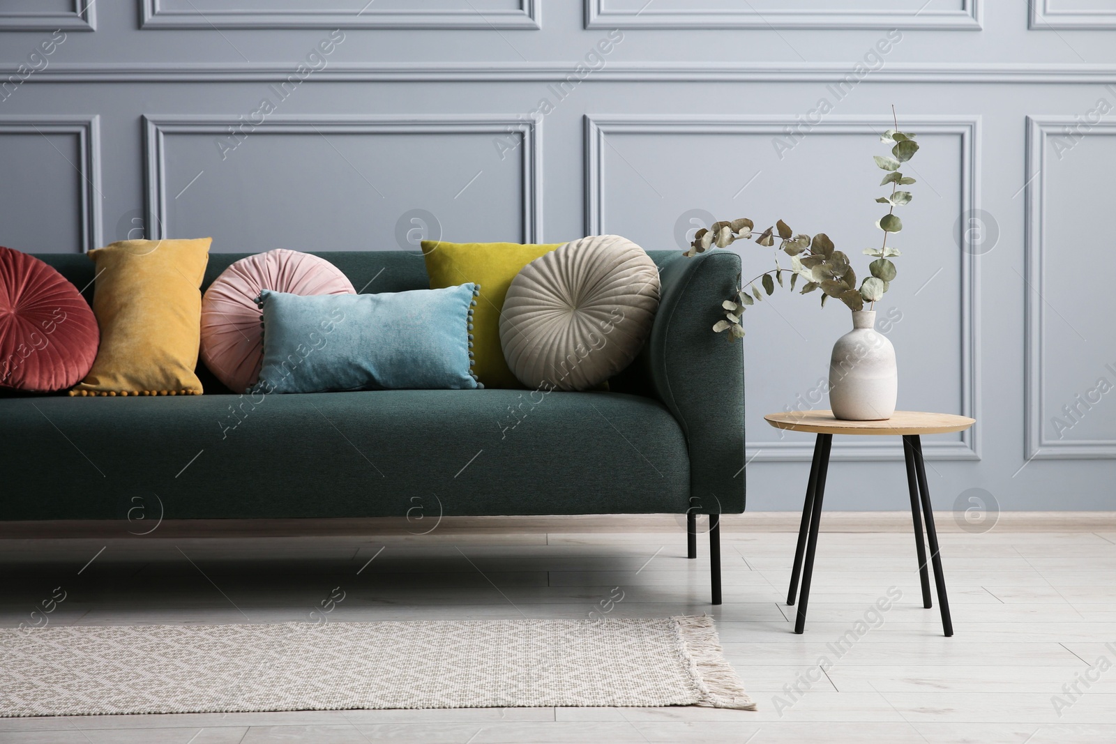 Photo of Stylish sofa with cushions and eucalyptus branches in vase on side table near grey wall