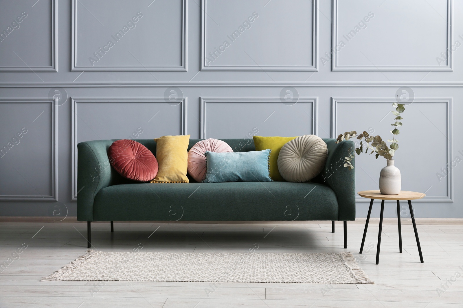 Photo of Stylish sofa with cushions and eucalyptus branches in vase on side table near grey wall