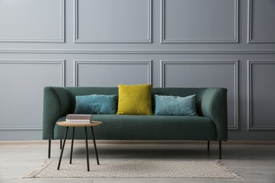 Photo of Stylish sofa with cushions and books on side table near grey wall