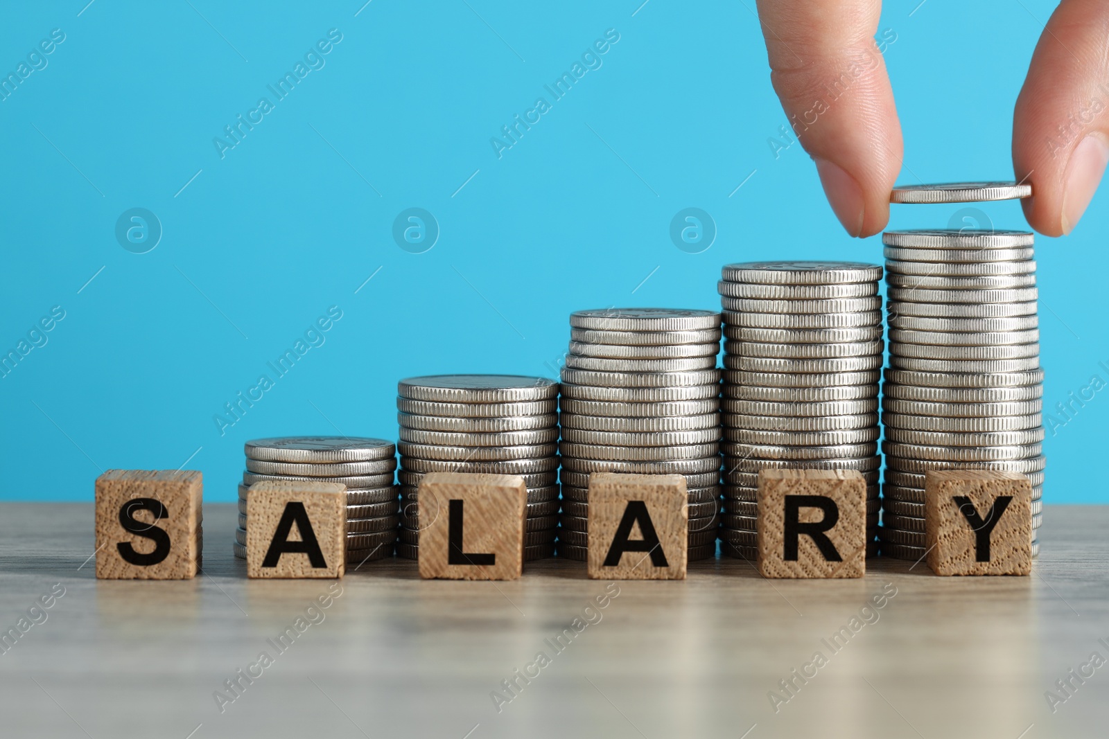 Photo of Woman putting coins and cubes with word Salary at wooden table against light blue background, closeup