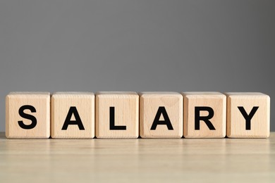 Photo of Word Salary made of cubes on light wooden table against grey background, closeup