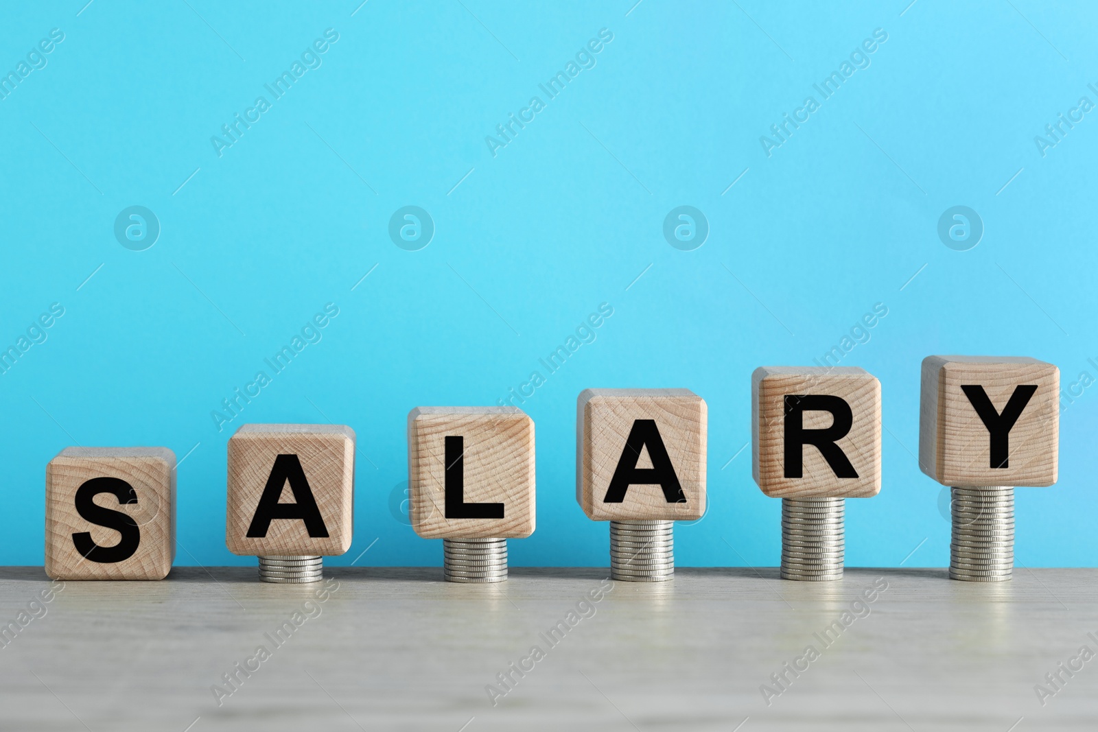 Photo of Word Salary made of cubes and stacked coins on wooden table against light blue background