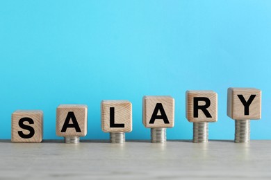 Photo of Word Salary made of cubes and stacked coins on wooden table against light blue background