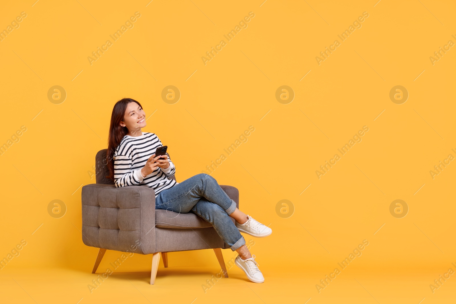 Photo of Smiling woman with smartphone sitting in armchair on yellow background. Space for text