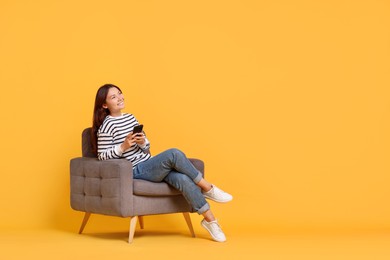 Smiling woman with smartphone sitting in armchair on yellow background. Space for text