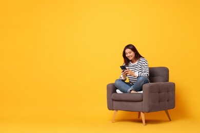 Smiling woman with smartphone sitting in armchair on yellow background. Space for text