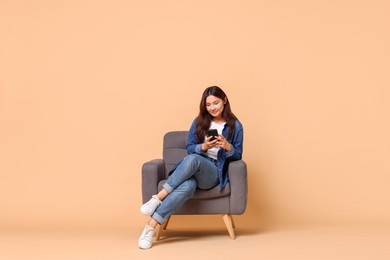 Photo of Young woman with smartphone sitting in armchair on beige background. Space for text