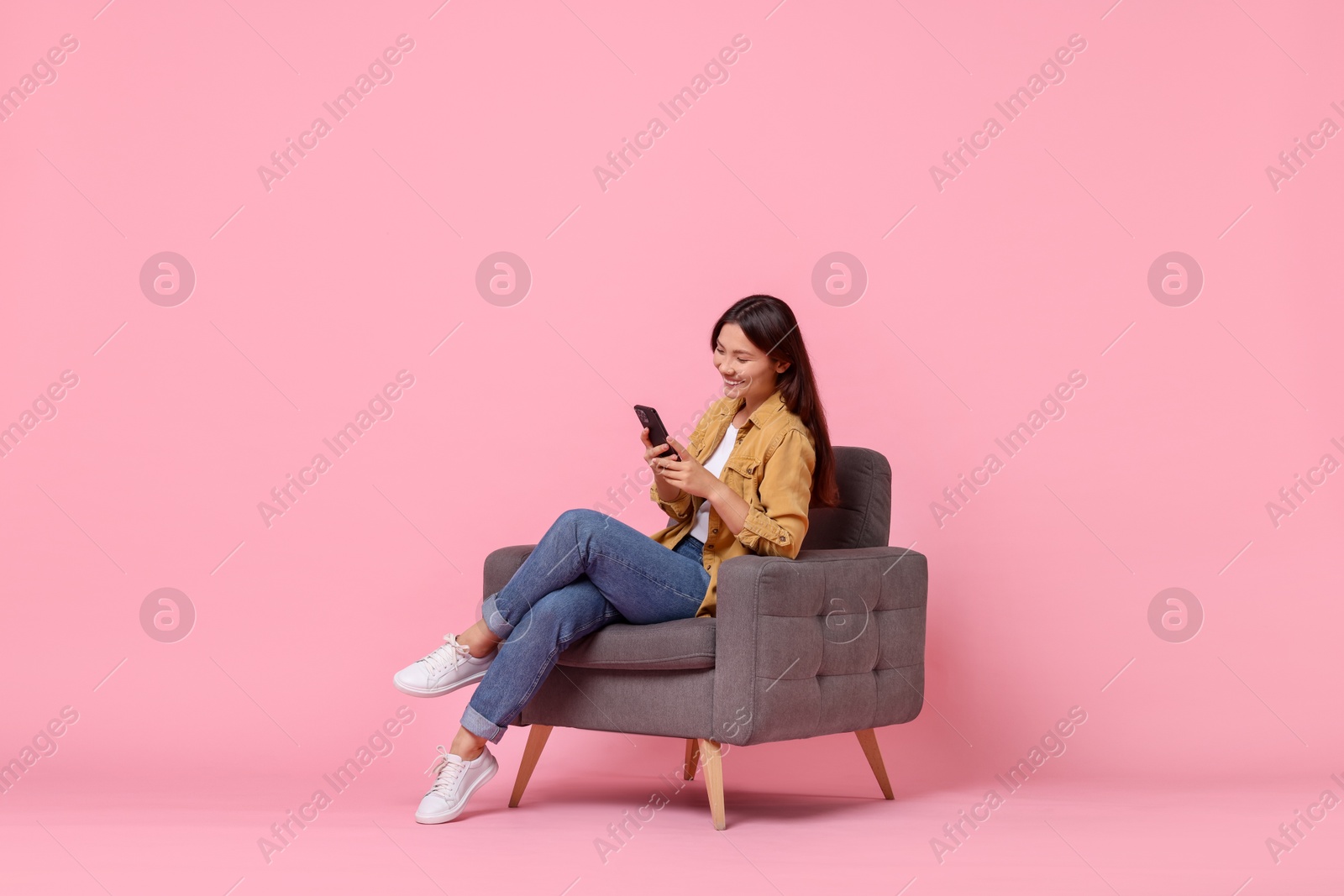 Photo of Smiling woman with smartphone sitting in armchair on pink background. Space for text