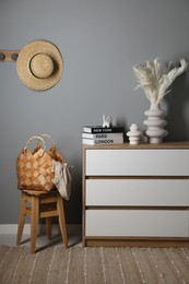 Different decorative elements on chest of drawers, chair and wicker bag near gray wall indoors