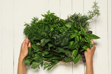 Photo of Woman holding different fresh herbs in basket at white wooden table, top view