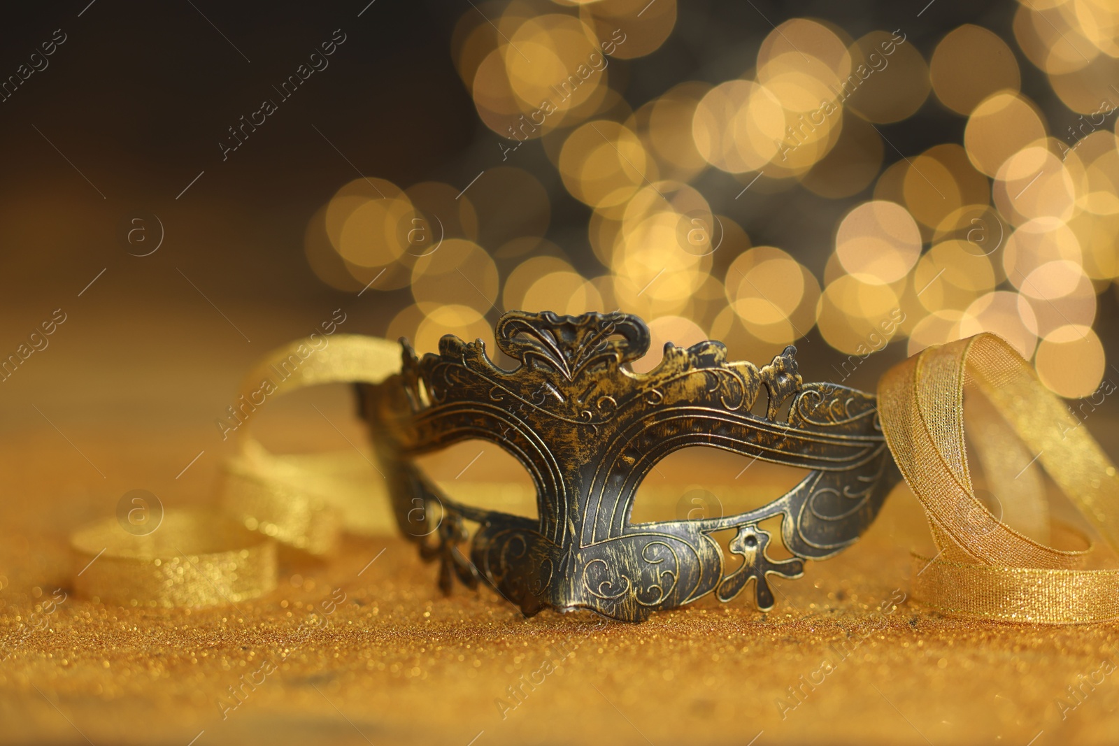 Photo of Beautiful carnival mask on golden shiny surface against blurred lights, closeup