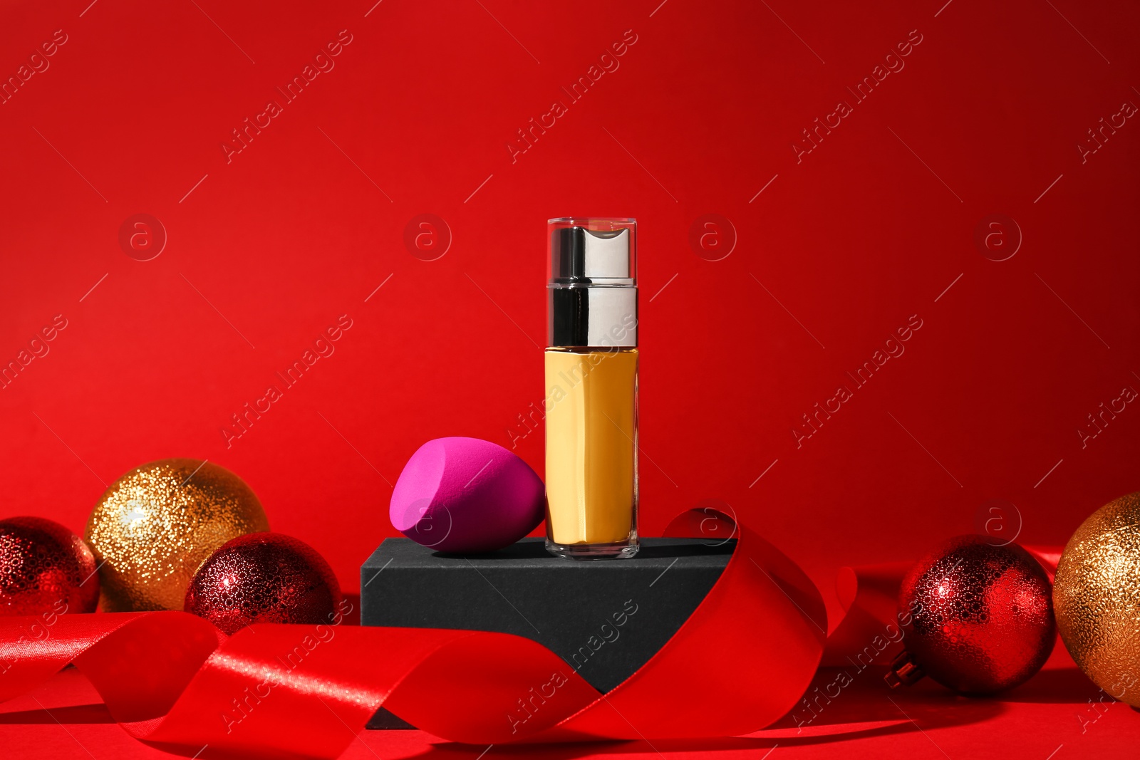 Photo of Christmas present. Liquid foundation, sponge, gift box and festive decor on red background