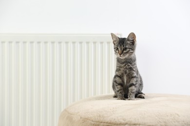 Photo of Cute little kitten on pouf near radiator at home, space for text