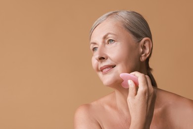 Photo of Beautiful woman doing facial massage with gua sha tool on beige background, space for text